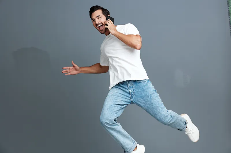 Happy smiling young man talking on smartphone while jumping in the air, dressed in casual white t-shirt and blue jeans against a simple gray background. Lifestyle concept.