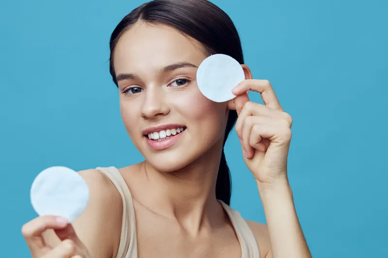 Young woman demonstrating skincare routine with cotton pads on a blue background, smiling and relaxed, highlighting beauty and self care concepts