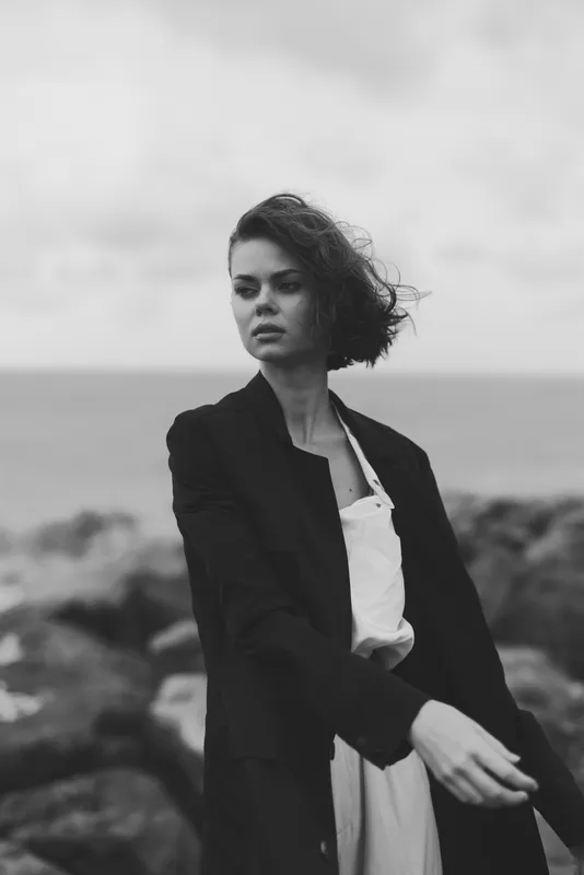 Monochrome image of a confident woman in a blazer by the seaside, wind in hair, thoughtful gaze, wearing a light dress, moody fashion photography with coastal backdrop and natural textures