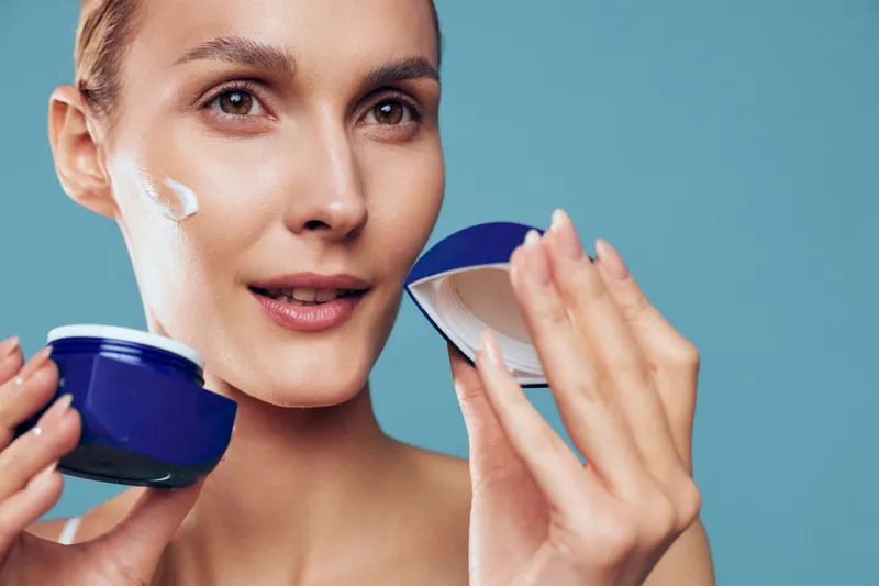 Young woman applies moisturizing cream on face holds open jar with cosmetic product looks at camera has natural skin care routine isolated on blue background.