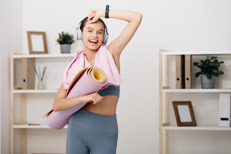 Woman fitness model warming up at home with a yoga mat, towel over shoulder, and headphones. Energetic workout scene highlighting sport, cardio, focus, and movement in a bright, tidy room.