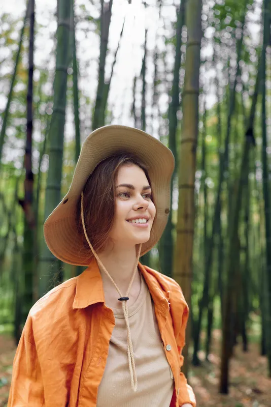 Woman hat bamboo forest smile portrait outdoors nature. Happy female traveler in orange jacket and beige top enjoying a peaceful green grove, relaxed and curious expression in daylight.