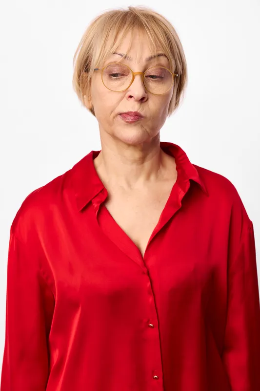 Woman portrait in studio, blond hair and glasses, red shirt, neutral white background, mature calm expression, gentle downward gaze, soft light