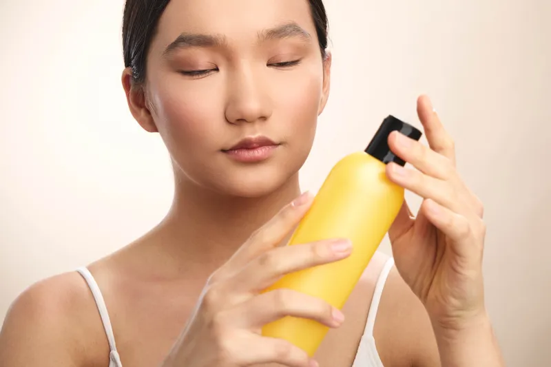 Asian woman gently applies moisturizer from a yellow bottle in a bright, clean bathroom, portraying a calm skincare routine and focused selfcare moment.