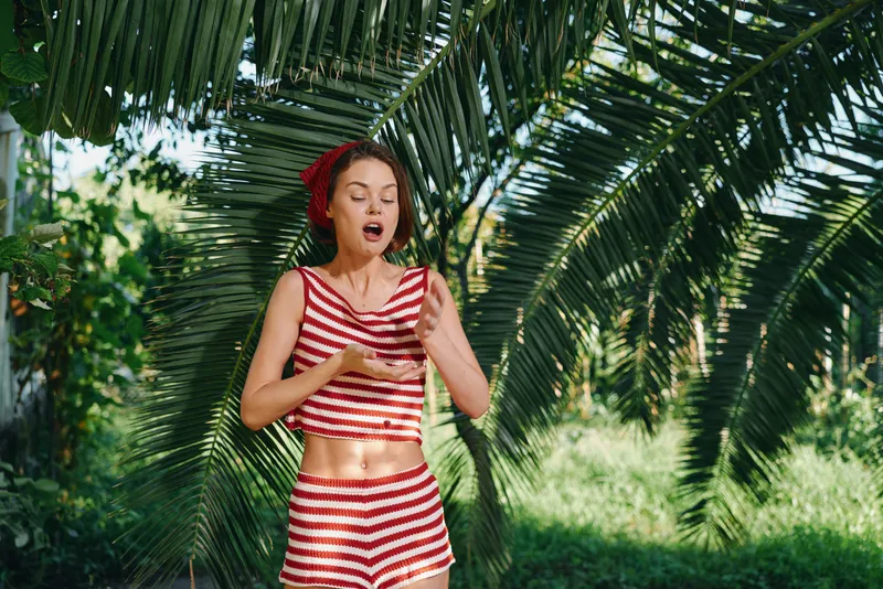 A cheerful woman wearing a red and white striped crop top and shorts stands beneath lush palm fronds in a sunny outdoor setting, expressing surprise with a playful, carefree mood.