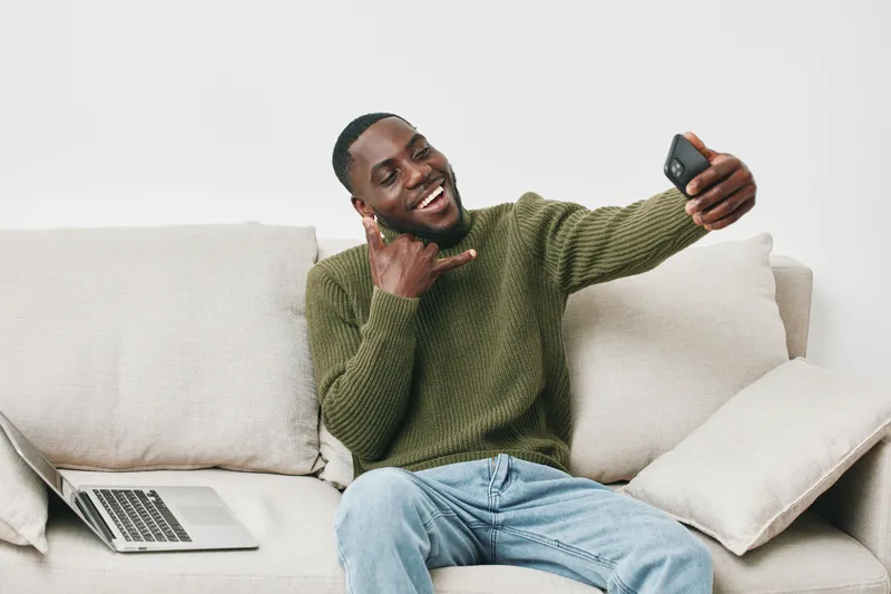Cheerful young Black man taking a selfie while sitting on a cozy sofa, wearing a green sweater, showcasing a relaxed and joyful emotion, perfect for lifestyle images