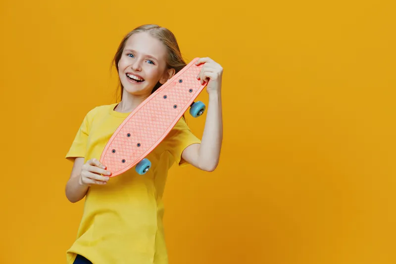 Happy girl with a pink skateboard on a bright yellow background, showcasing youthful energy and joy in a playful summer scene
