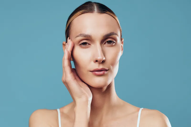 Close-up portrait of a young woman with clear skin touching her face on a blue background. Concept of skincare, natural beauty, and facial care with soft studio lighting.