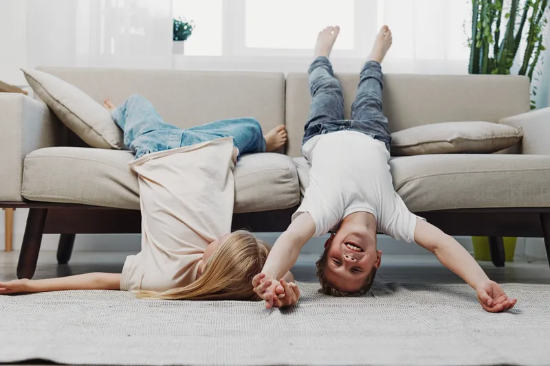 Two happy children lie upside down on the sofa holding hands in a bright living room, having fun and enjoying playful moments together in a cozy home environment.