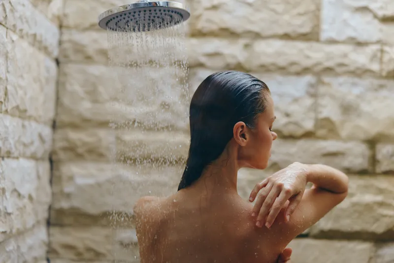 woman showering outdoors with wet hair under rainfall showerhead, natural stone wall background, skincare routine, fresh and relaxing moment