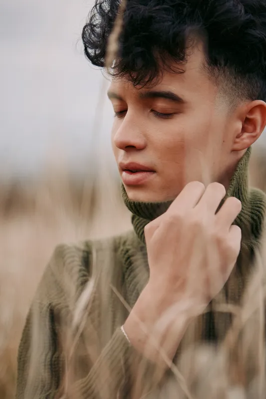 A close portrait of a young man in a turtleneck sweater standing in tall dry grasses, eyes gently closed, capturing quiet emotion, natural light and authenticity with understated, accurate feeling.