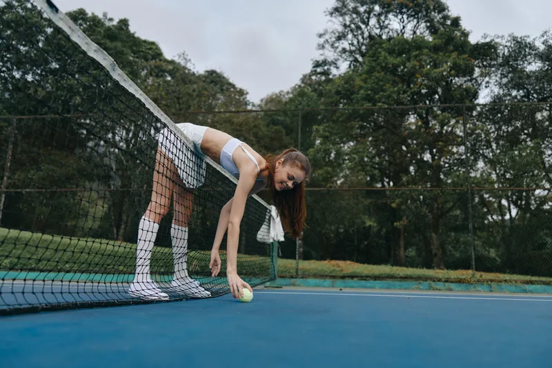 A young woman in sportswear picking up a tennis ball on a court surrounded by greenery, showcasing focus and determination for an active lifestyle