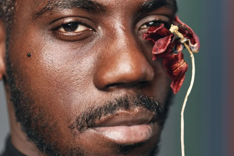 African man portrait with dark skin and facial hair holding dried red flower near eye in creative conceptual visual style closeup with textured skin and serious expression.