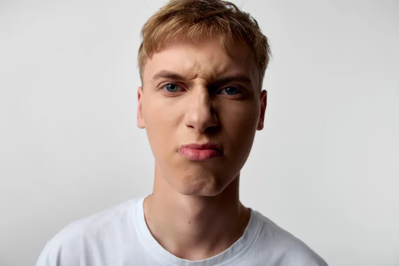 Close up of an annoyed man with angry facial expression, pursed lips, and frustrated gaze, wearing a white t shirt against a plain backdrop, conveying irritation, mood, and intense emotion suitable