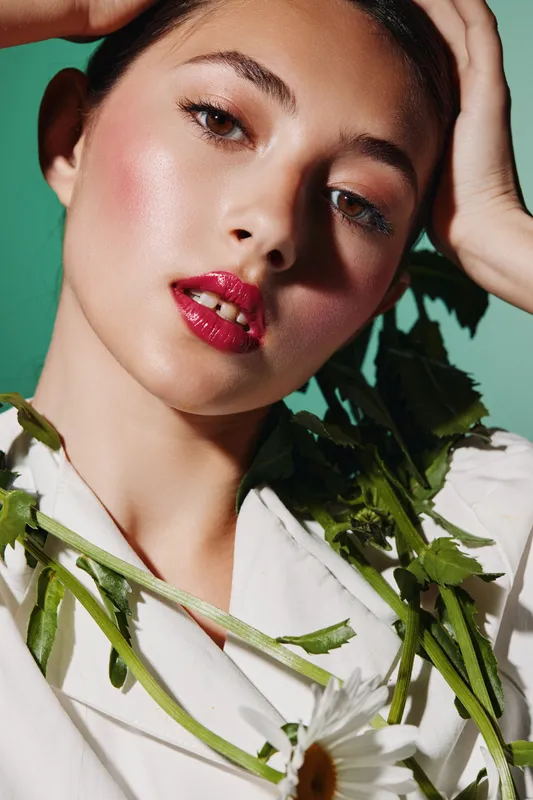 Close-up of a young woman with vibrant red lipstick and dewy skin against green. She wears white with greenery, showcasing beauty, fashion, and creative style.