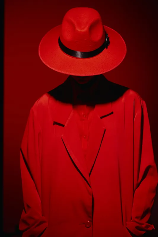 Fashion model wearing vivid red hat and matching oversized jacket stands against deep red backdrop. Dramatic shadow hides facial features, creating intense beauty and style concept.