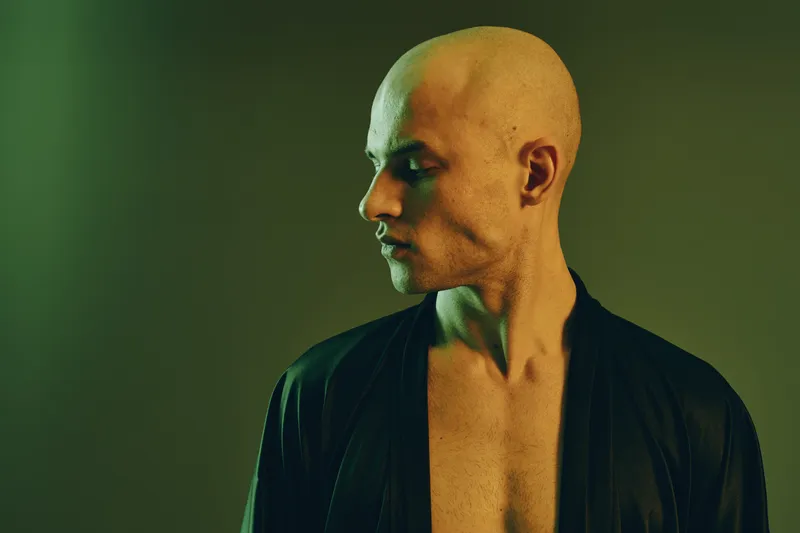 Portrait of a man with a shaved head, demonstrating calm expression against a light green background. Captivating lighting adds depth to the serene atmosphere.