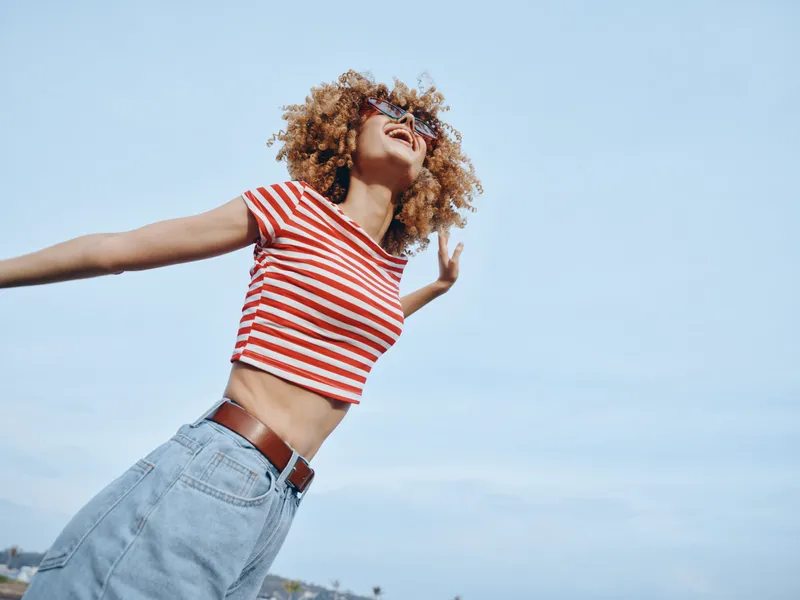 Joyful woman with curly hair in a striped crop top and jeans dances outdoors under a clear blue sky, capturing carefree energy and spontaneous movement.