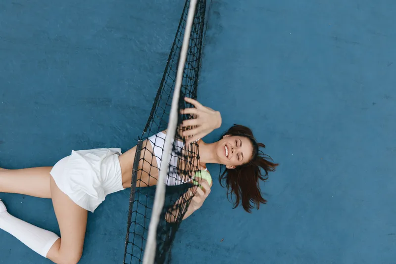Young woman enjoying tennis on court, wearing sportswear, smiling with tennis ball in hand, vibrant blue background, perfect for sports and fitness themes