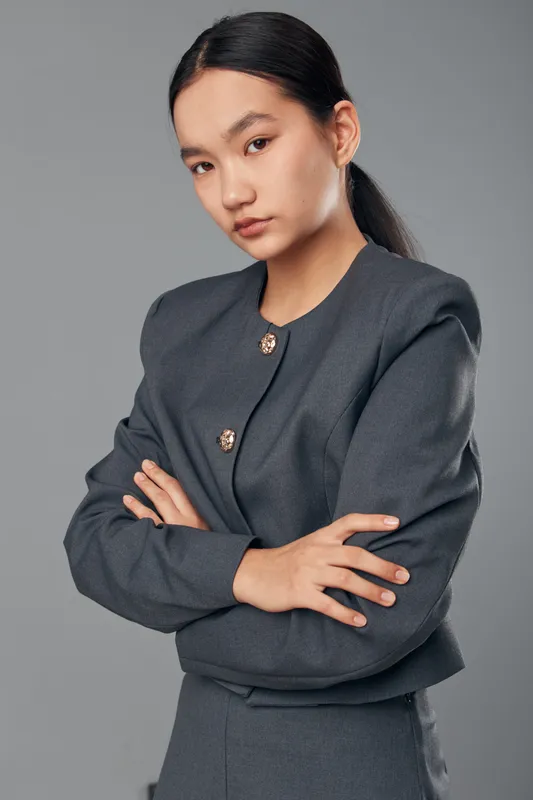 Young woman with serious expression posing with crossed arms in formal gray suit on neutral background. Professional, confident model with straight black hair and natural makeup in studio.