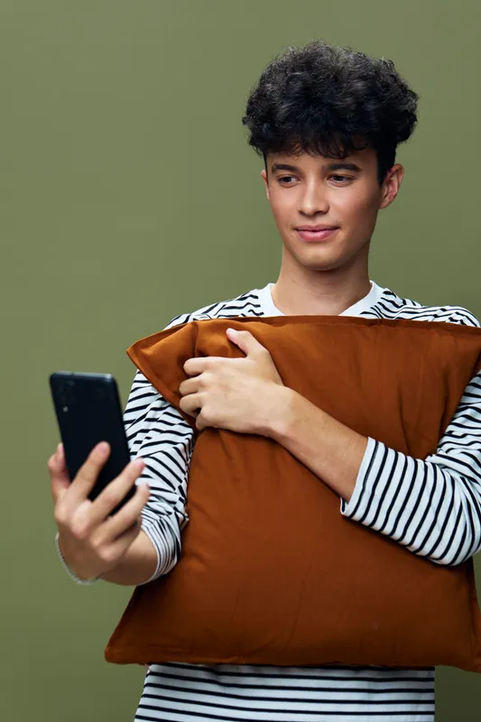 young man holding brown pillow and taking selfie with smartphone wearing striped shirt against green background in cozy indoor setting