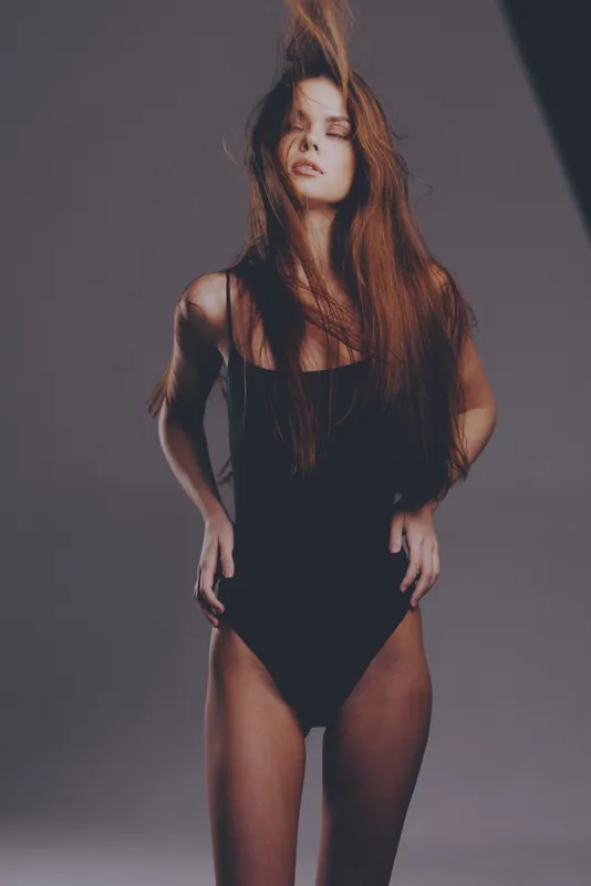 Young woman in black bodysuit posing with her hair flowing against a neutral background, expressing confidence and movement.
