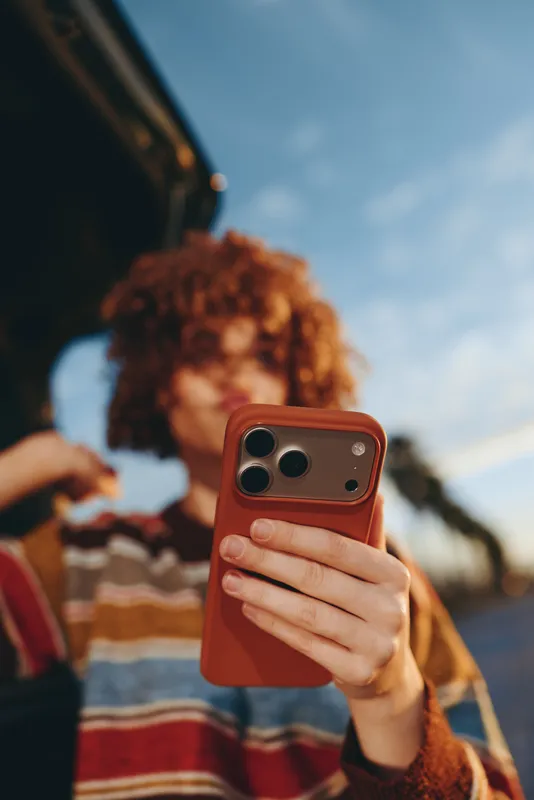 Woman lifestyle selfie moment outdoors, curly hair visible, holding mobile smartphone and red phone case, portrait framing, casual striped sweater, soft daylight, blurred people background, candid