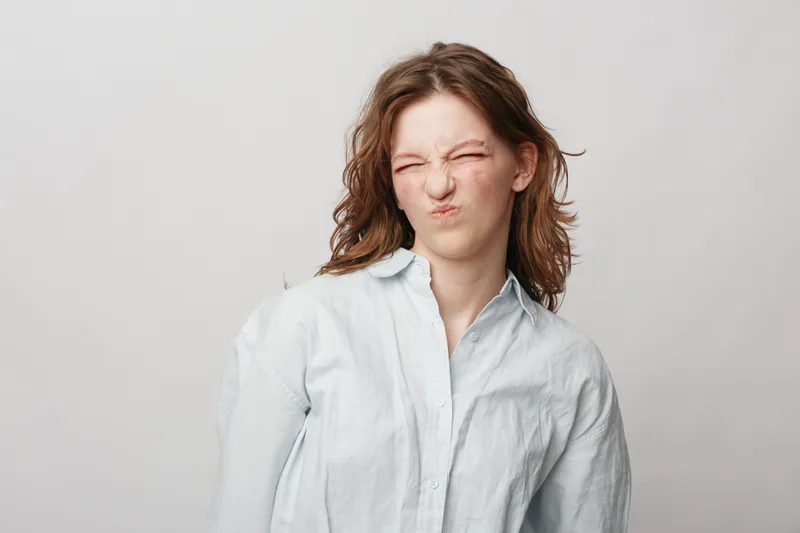 Young woman with brown hair wearing white shirt makes a funny face with eyes closed and lips scrunched. Isolated on light background. Concept of humor, emotion, and expression.