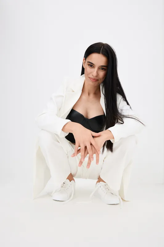Young woman crouching confidently in white suit and black top with long dark hair and white sneakers against plain white background, modern casual style.