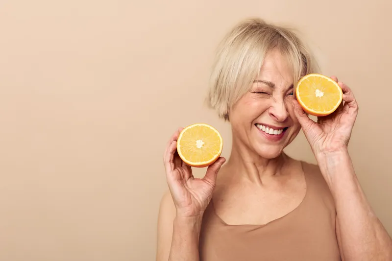 Happy mature woman with short blonde hair holds two orange halves near face and smiles with eyes closed on beige background. Concept of healthy lifestyle, vitamin c, and natural skincare.