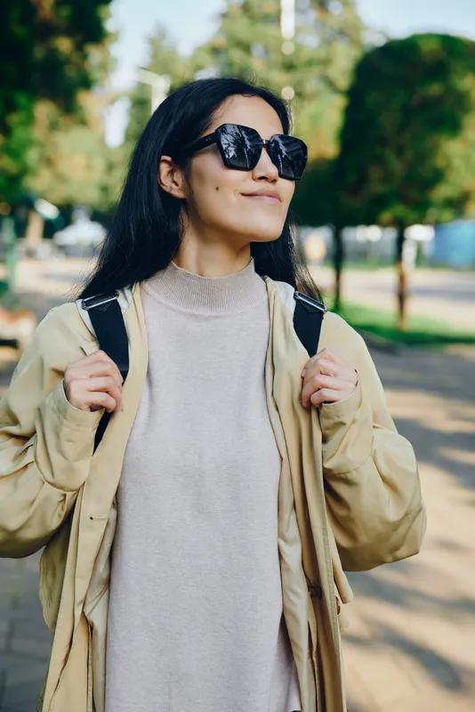 Woman wearing sunglasses, beige coat and knitted sweater, backpack over shoulders, standing in a sunny park with street vibes, fashion forward and confident lifestyle image