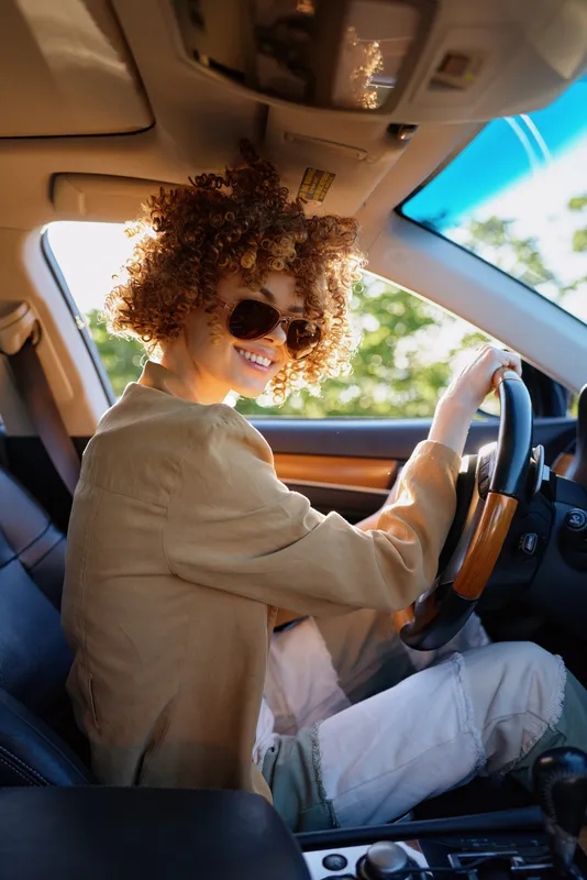 A driver with curly hair wears sunglasses and smiles while seated in a sunlit car, showcasing a casual lifestyle with retro film color and warm, natural tones inside.