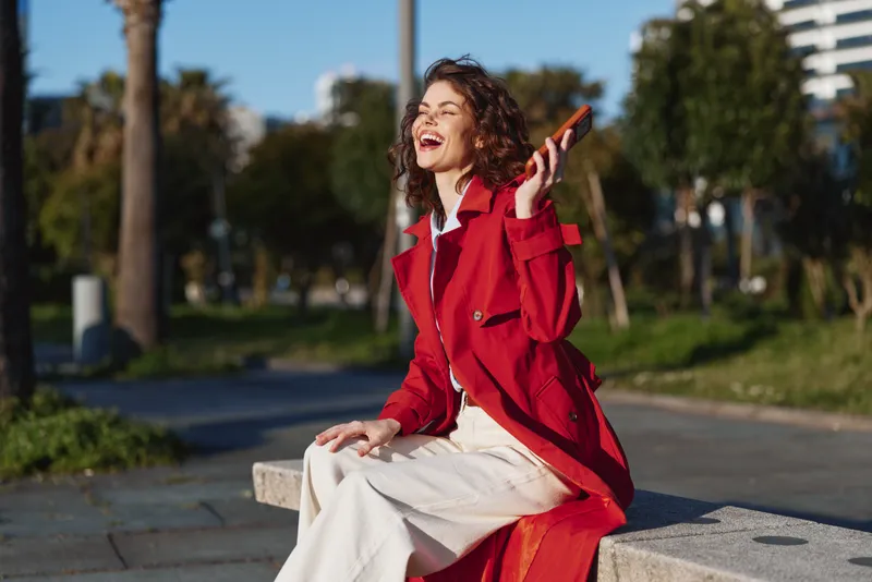 Smiling woman in a red coat sits on a concrete bench in a sunny park, talking on a smartphone. Fashionable street style, casual elegance, joyful outdoor moment.