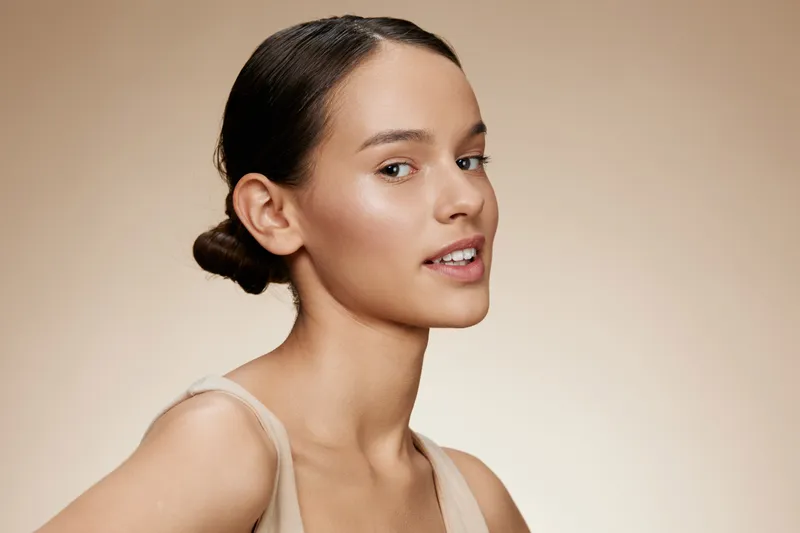 A serene young woman with natural makeup poses against a soft beige background, showcasing healthy skin and an elegant hairstyle, embodying beauty and confidence