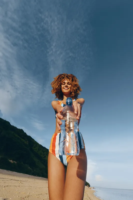 Woman on beach holding water bottle, summer sunlight, curly hair blowing, smiling gaze upward, clear blue sky, ocean breeze, scenic coastline, bright colors, relaxed pose