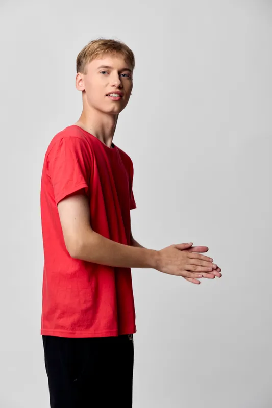 Hands, clapping and a young male in a red shirt stand against a neutral studio backdrop, conveying celebration, approval, or readiness for action in a casual promotional scene.