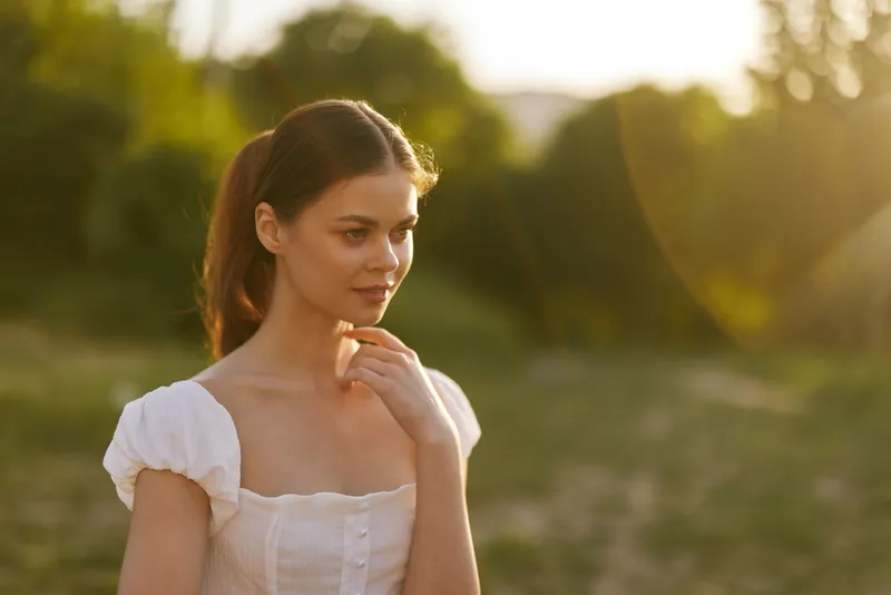 Thoughtful young woman in a white dress enjoying a sunny day outdoors, surrounded by nature, radiating joy and serenity