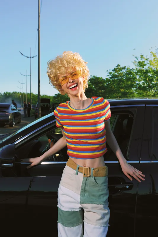 Cheerful woman outdoors, smiling beside a dark car, wearing a striped rainbow top and colorblock shorts, sunlit breeze, casual lifestyle, vivid retro colors, joyful moment, warm light, candid pose,