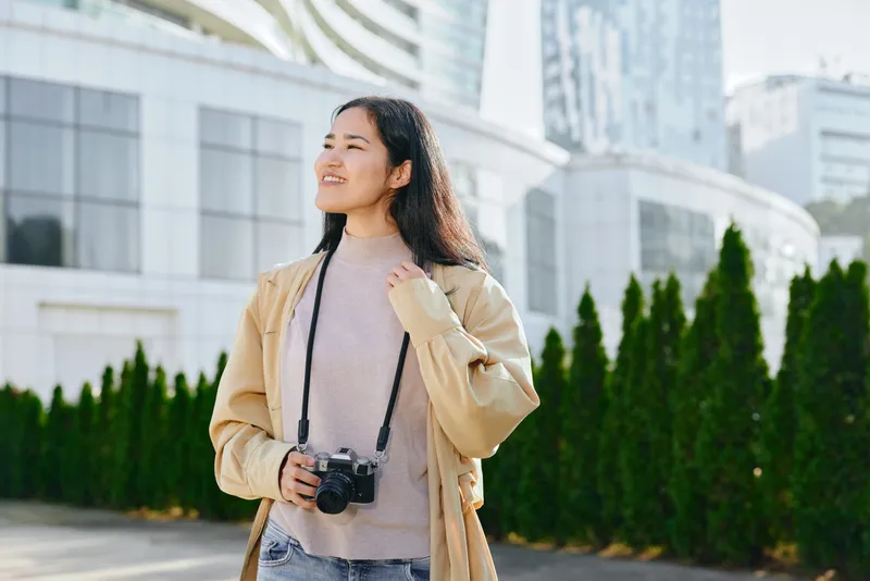 Camera in hand, a woman explores an outdoor urban setting, smiling beside modern architecture and green hedges, wearing a beige jacket and soft sweater, capturing a lifestyle travel moment
