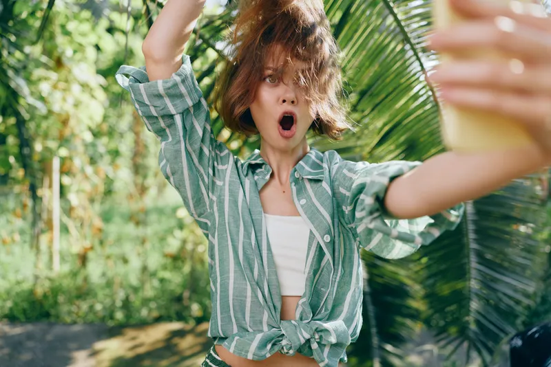 A young woman in a green striped shirt tied at the waist stands outdoors with her arms raised, surprised expression under bright sun.