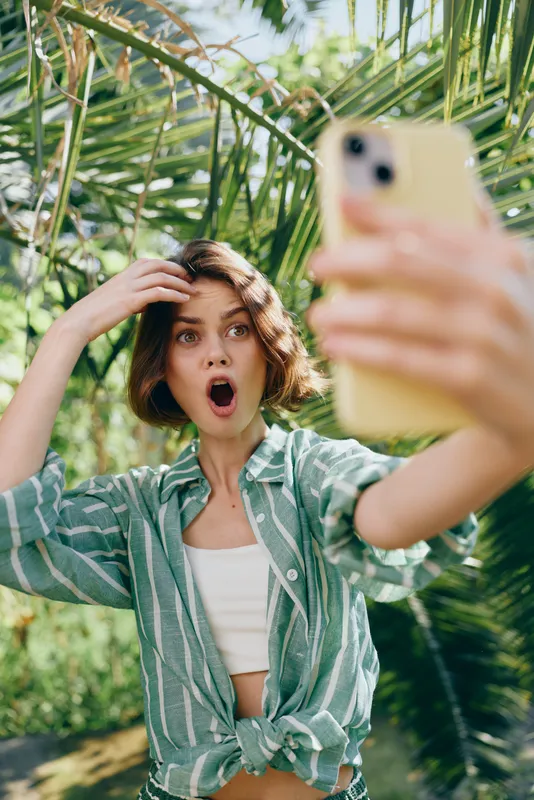 Young woman in a green striped shirt takes a surprised selfie in a lush garden, sunlight filtering through leaves, capturing a playful, spontaneous moment.