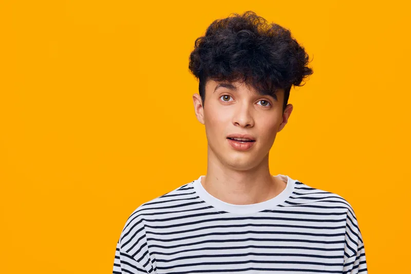 young man with curly hair wearing striped shirt looking surprised, expressive face, bright orange background, casual style, modern portrait