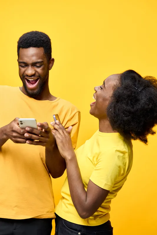 Joyful young couple in matching yellow outfits laughing at a smartphone against a bright yellow background, showcasing happiness and connection