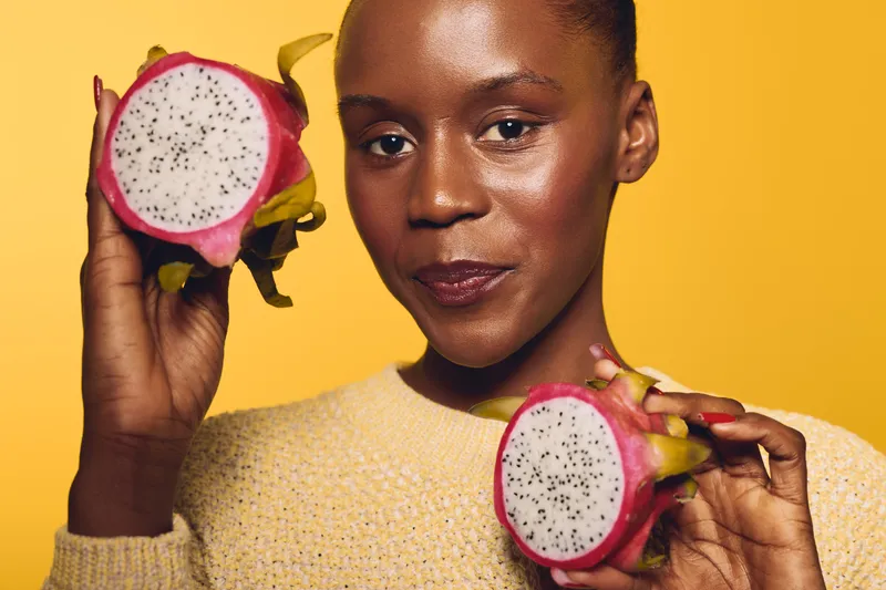 Close-up of a smiling dark skinned African woman holding two halves of a dragon fruit against a yellow background. Concept of healthy lifestyle, exotic fruit, and natural beauty.
