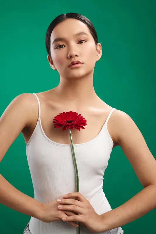 asian woman stands against a vivid green backdrop, holding a red flower at chest height, calm expression, graceful pose, soft lighting, contemporary portrait with a serene mood.