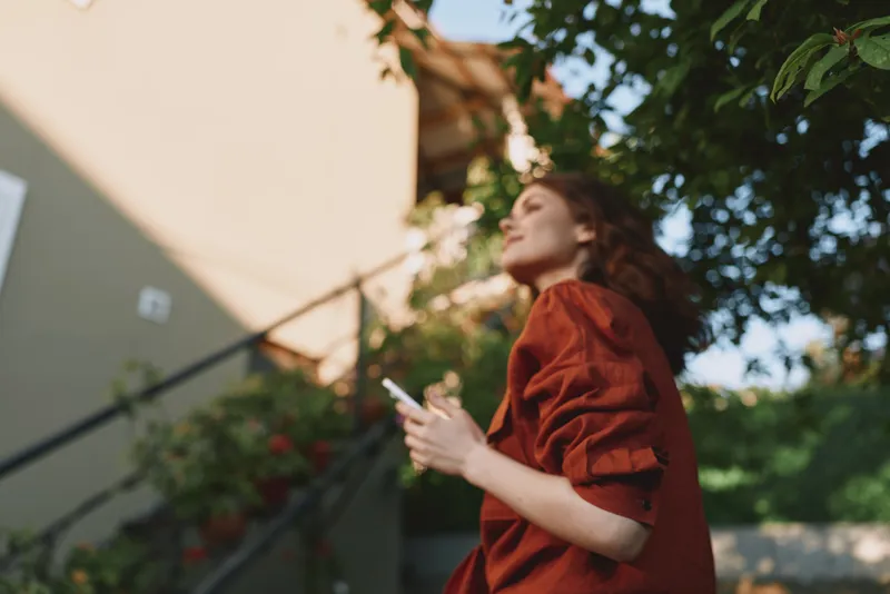 Young woman in a red shirt holding a smartphone outdoors, capturing a relaxed lifestyle moment under warm sunlight, with lush greenery in the background and soft, natural lighting, creating a casual