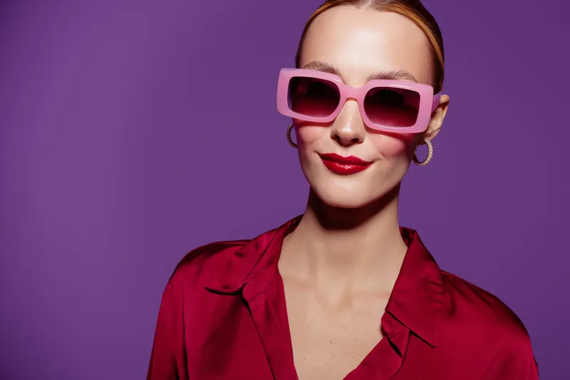 Stylish woman in pink sunglasses and red blouse poses confidently against purple background. Fashion portrait with bold colors and modern accessories for trendy lifestyle concept.