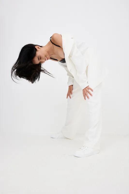 Woman in white suit bending forward with flowing black hair against plain white background in stylish contemporary fashion portrait with dynamic pose.