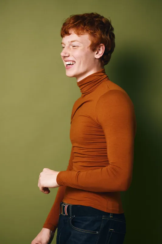 Young man with red hair laughing wearing orange turtleneck and dark pants standing in profile against green background indoors.