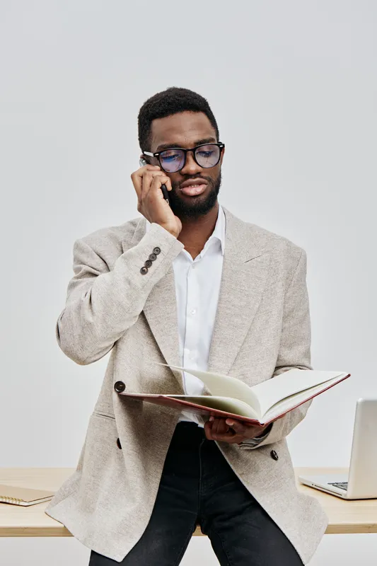 Confident professional young man with glasses and beard in beige blazer and white shirt, reading a book while talking on mobile phone, standing at office workspace with laptop and documents. Business 